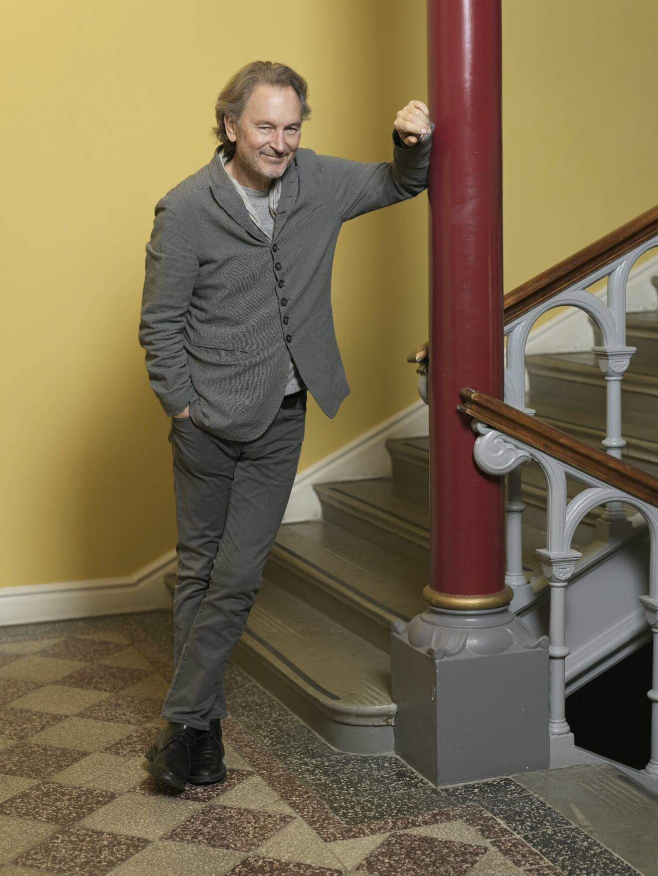Tomas Ledin i grå kostym, lutar sig mot en röd pelare i ett gult trapphus.
