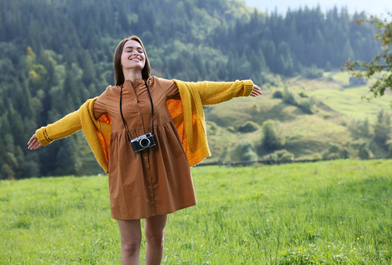 En ung kvinna med kamera runt halsen på en äng med berg i bakgrunden.