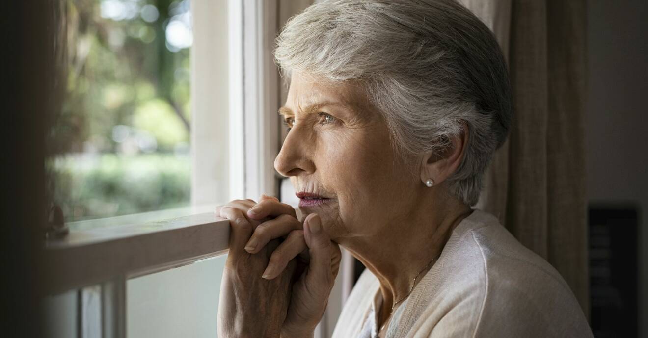 En äldre till synes deprimerad kvinna står och tittar ut genom ett fönster.