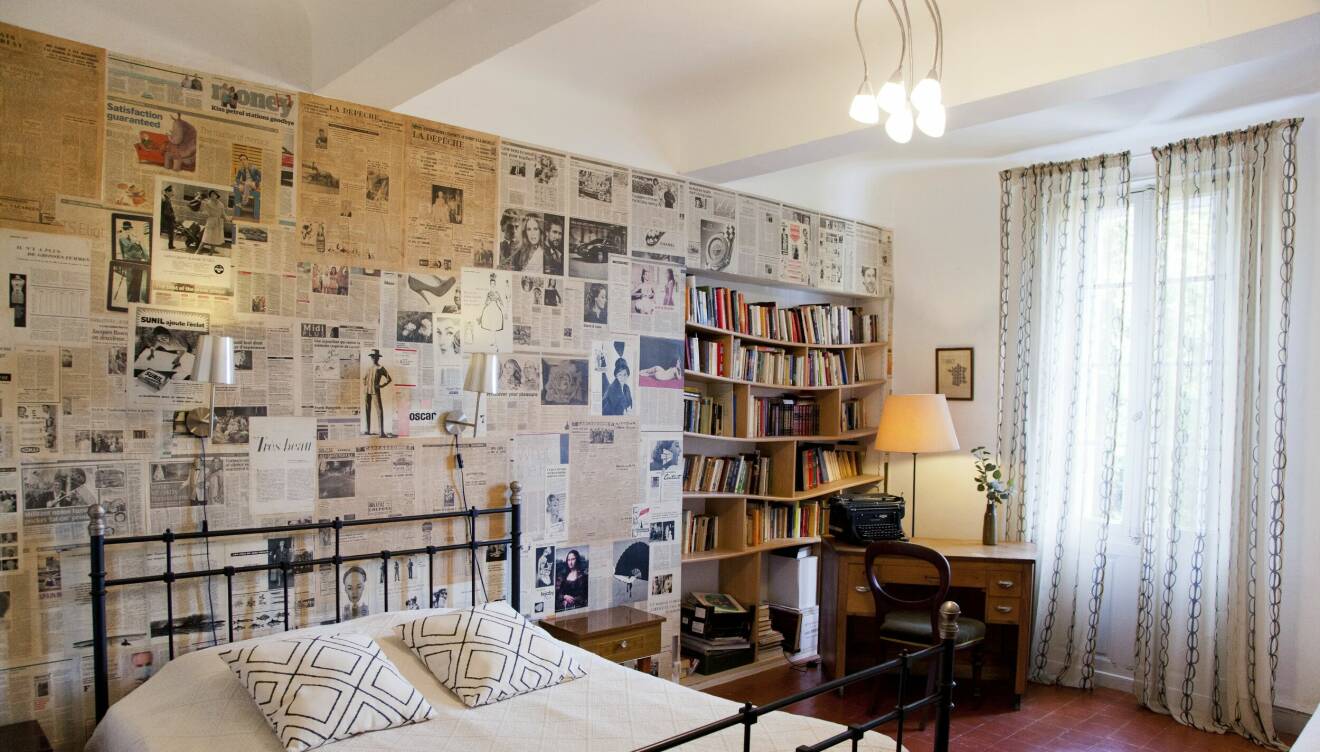 I Albert Camu-rummet är väggen vid huvudänden av sängen tapetserad med tidningsartiklar, bredvid sängen finns en inbyggd bokhylla med böcker och intill den står ett skrivbord.