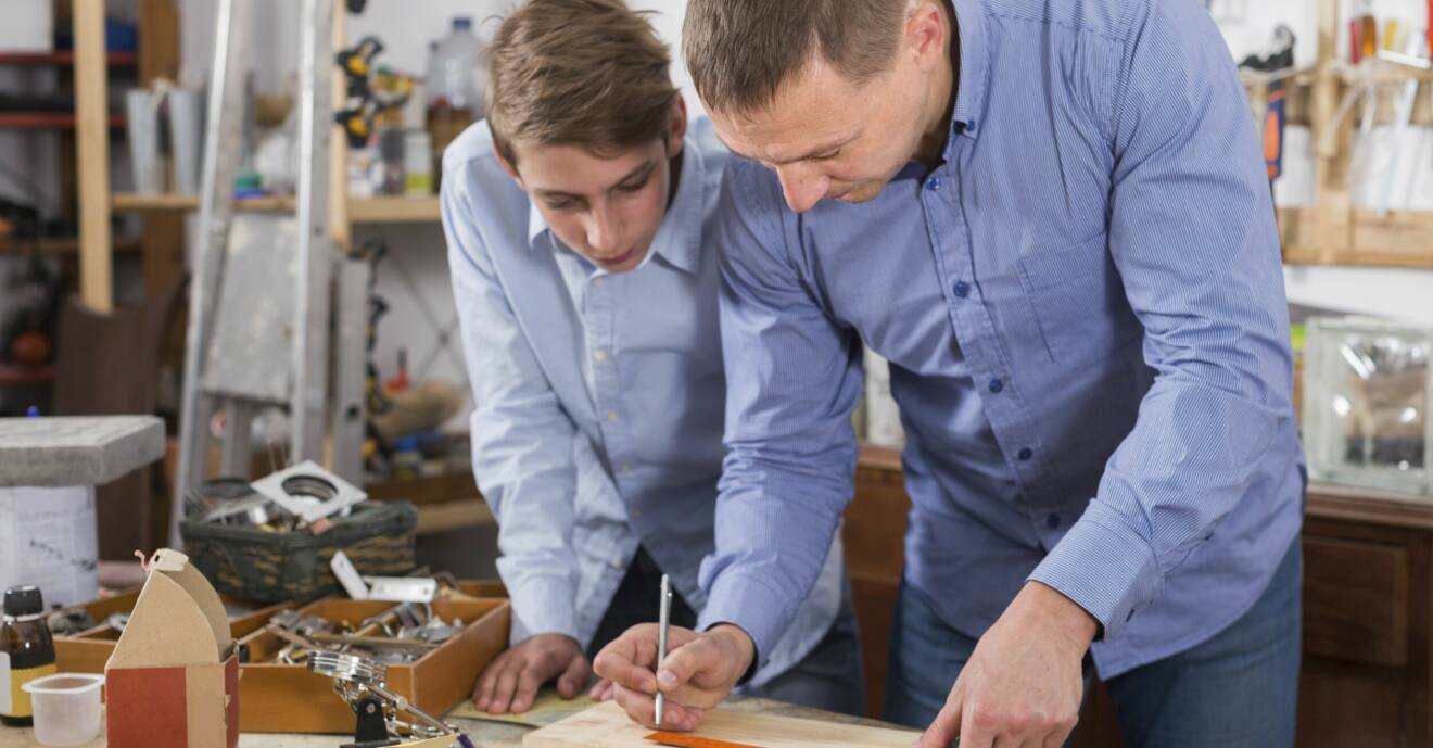 Pappa står i verkstaden med sin son och mäter och ritar på en bräda, lär demonstrerar hur man ska tänka när man snickrar.