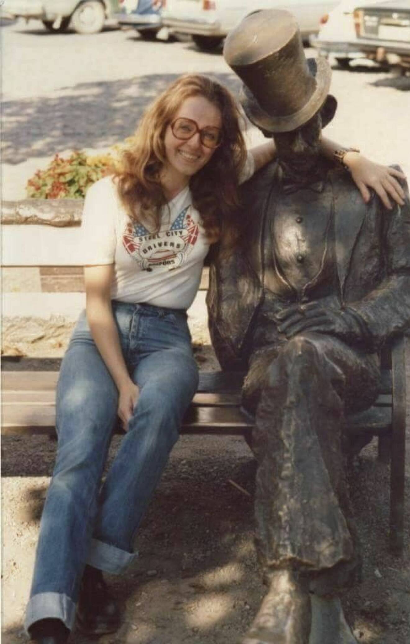 En ung Eva, iförd motorklubbtröja, sitter på en bänk i Filipstad, med armen om Nils Ferlin-statyn.