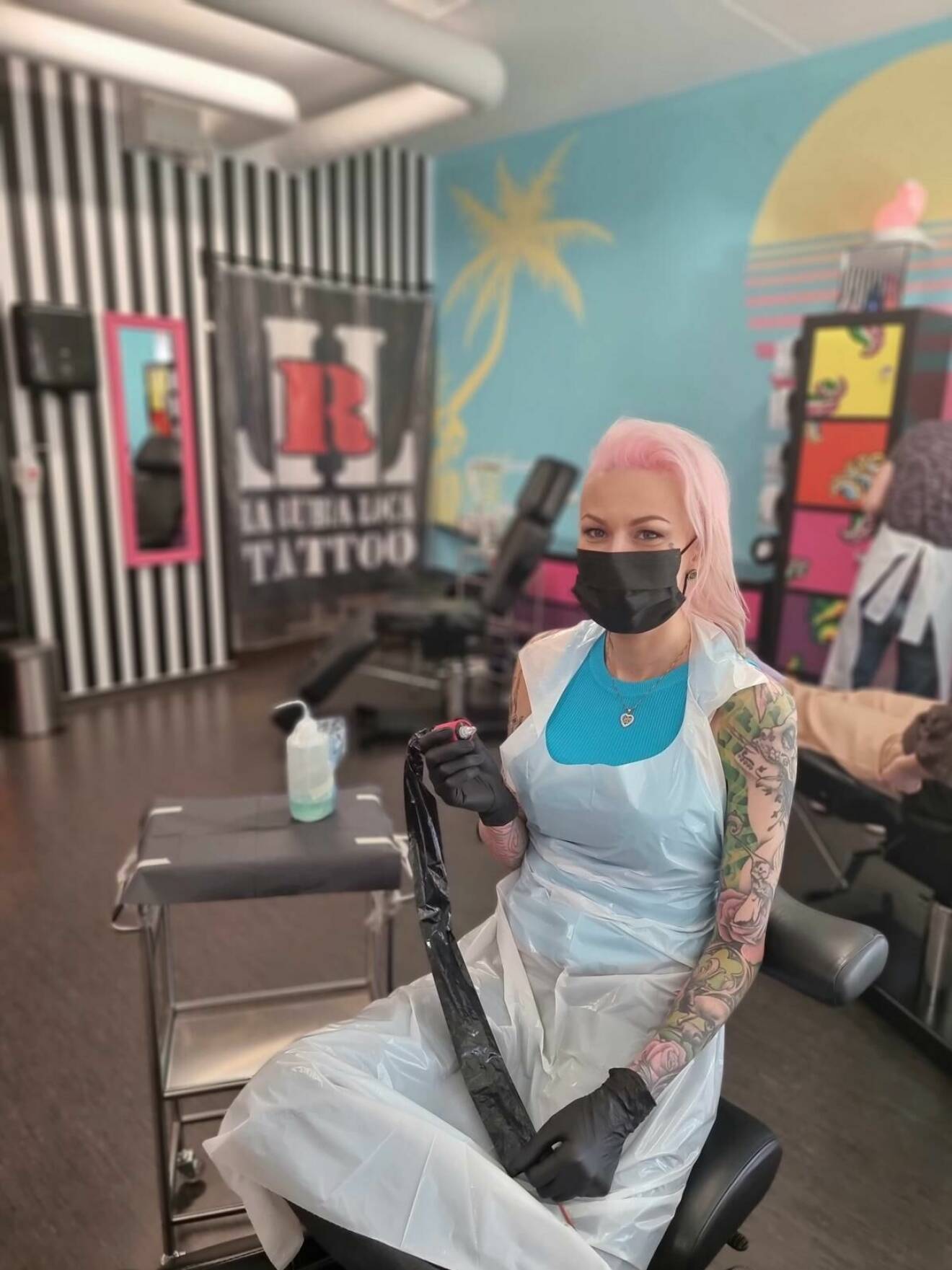En ung kvinna med rosa hår och munskydd håller i en tatueringsmaskin.