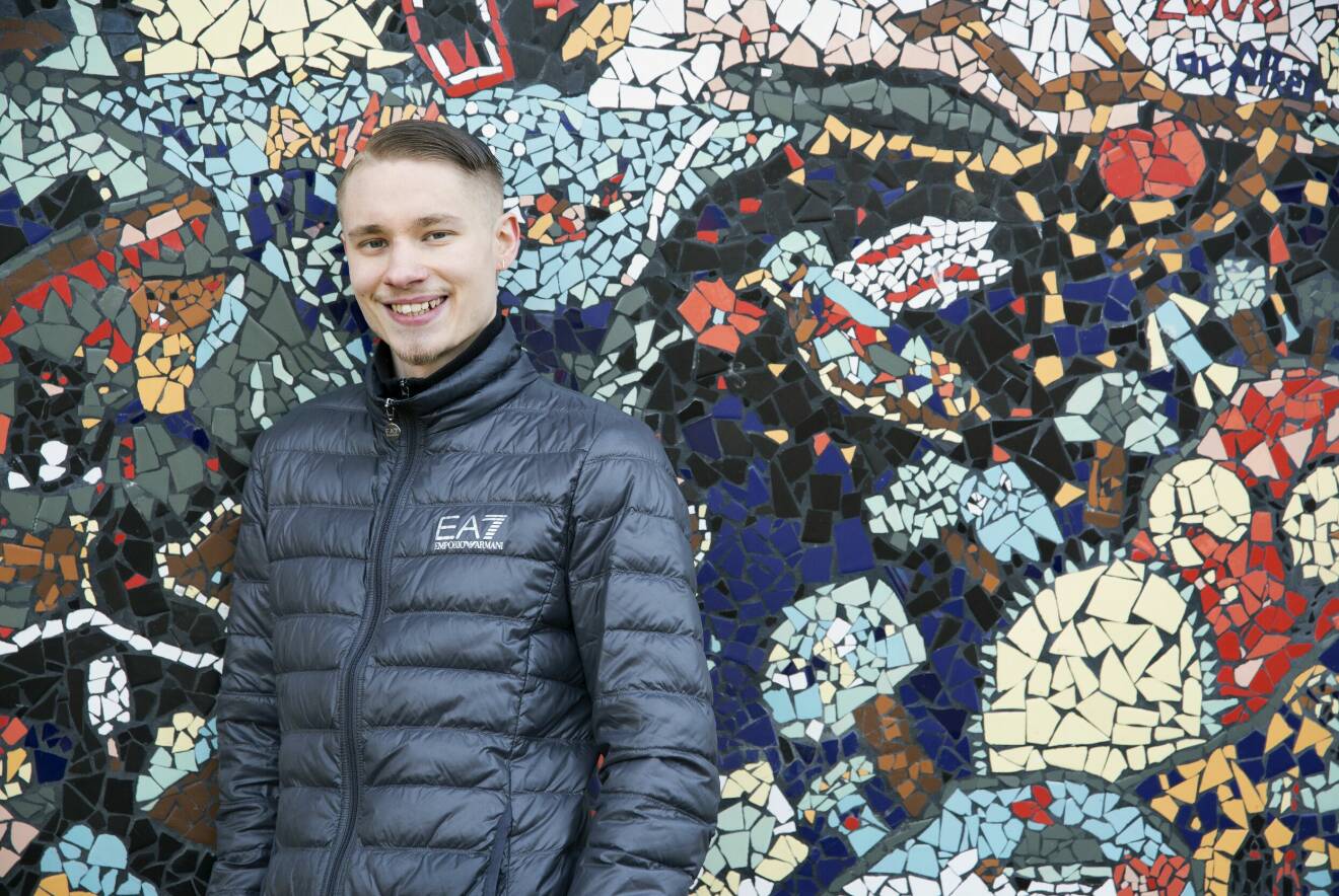 En ung man står leende framför en mosaikvägg.
