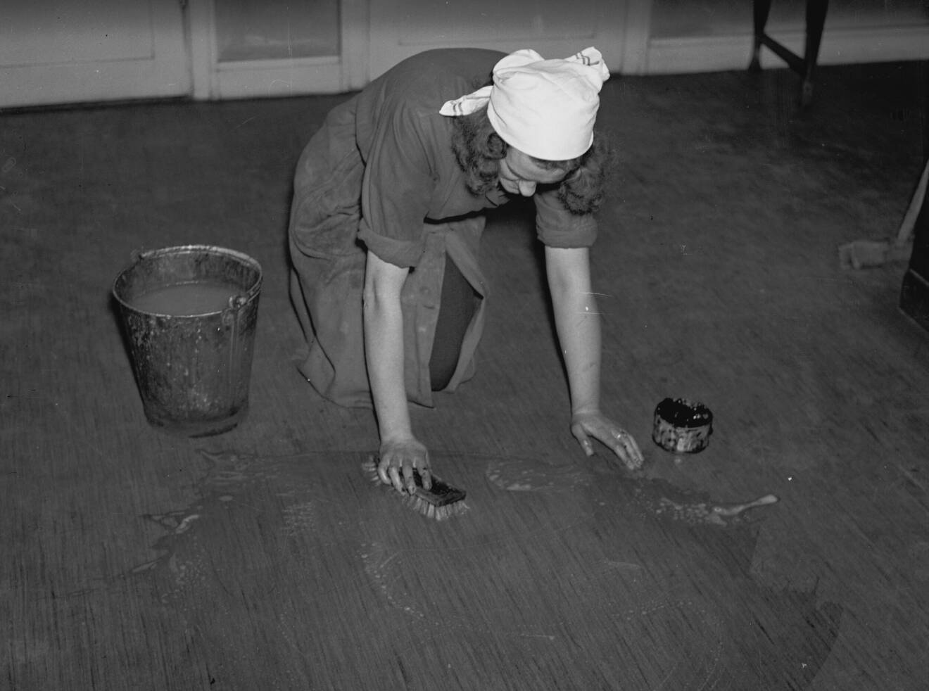 En städerska på 1970-talet, står på knä och skrubbar ett golv med en skurhink intill sig. Svartvit bild.