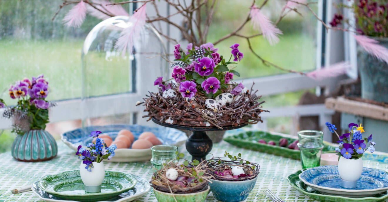 Dukat påskmiddagsbord med blomsterarrangemang i lila toner skapat kring ett fat på fot.