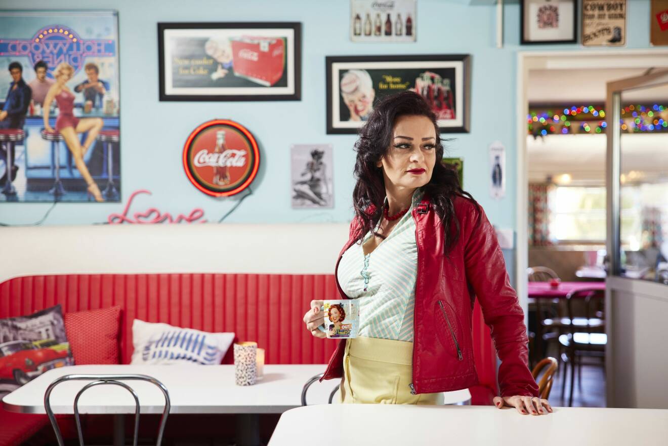 En medelålders kvinna i mörkt hår och röd skinnjacka står i ett 1950-talsinspirerat kök.