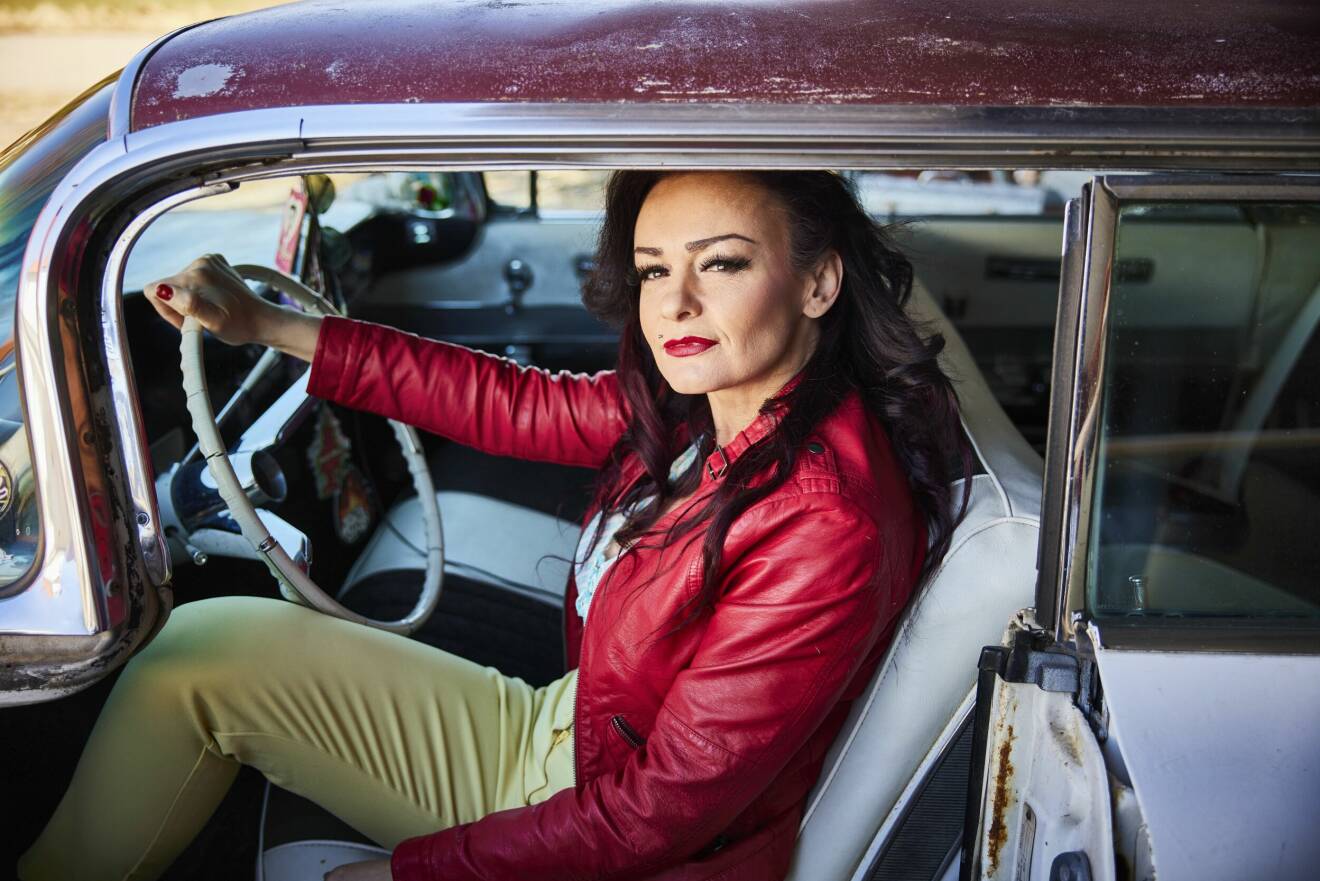 En medelålders kvinna i mörkt hår och röd skinnjacka sitter i en gammal bil från 1950-talet.