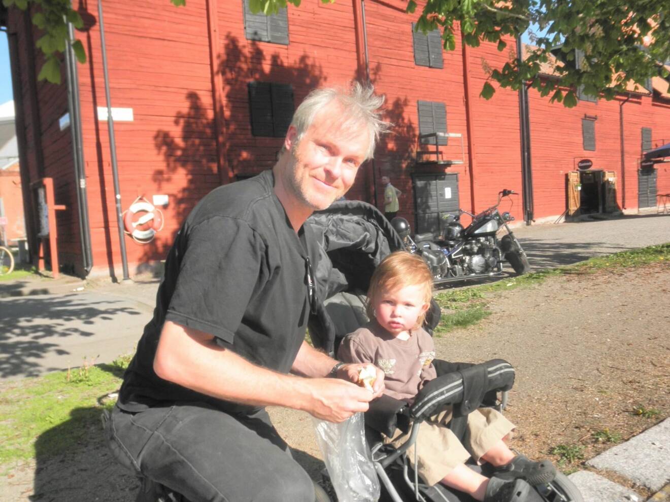 En man i 45-årsåldern sitter på huk framför en faluröd byggnad med sin lille son i barnvagn.
