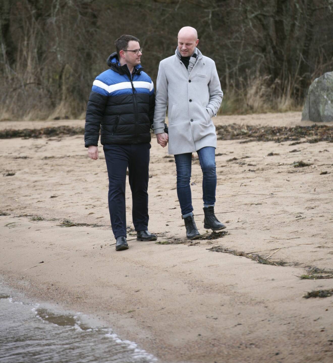 Johan och Mikael promenerar hand i hand vid en strand.