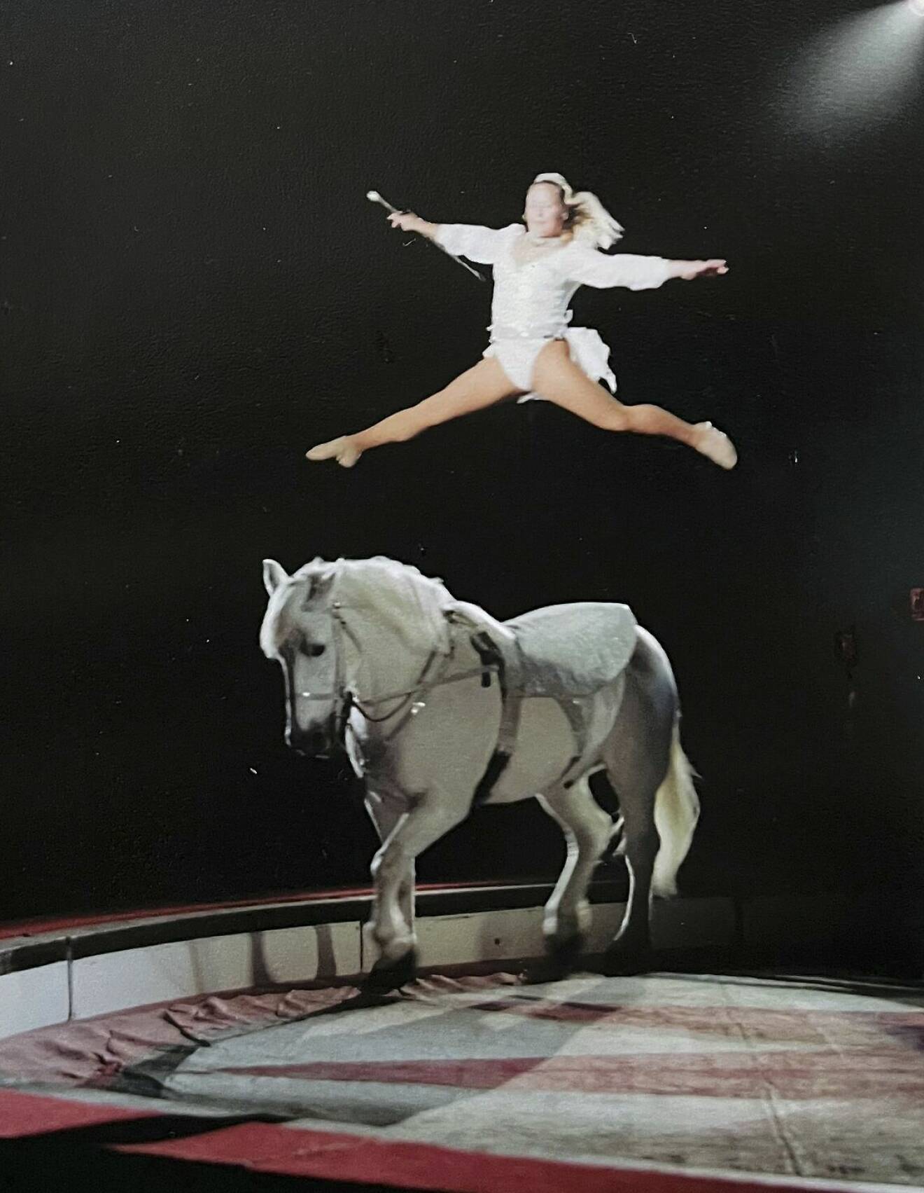 Susanne gör ett nummer med häst i manegen och har hoppat upp i luften och sträcker fram ena benet och sträcker det andra bakåt