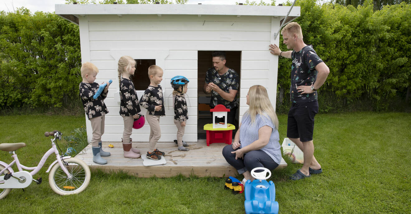 Hampus Engström sitter i en vitmålad lekstuga och leker med sina fyra barn som står uppradade utanför. Bredvid sitter Linda Fridland och Erik Fridland lutar sig mot lekhusets vägg.
