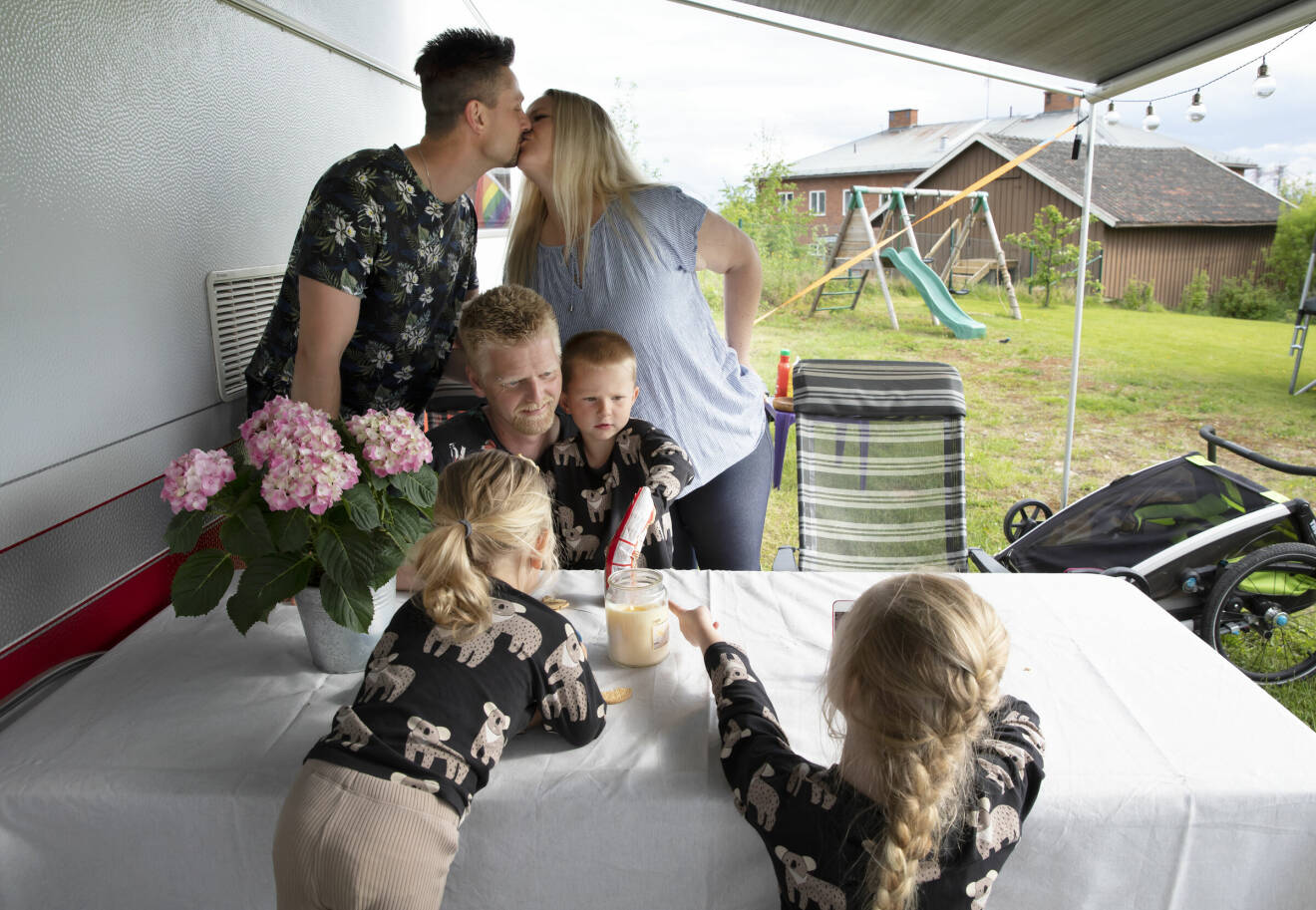 Vid ett bord intill en husvagn sitter Erik Fridland tillsammans med barnen Signe, Nils och Ture. I bakgrunden ger Hampus Engström och Linda Fridland varandra en puss.