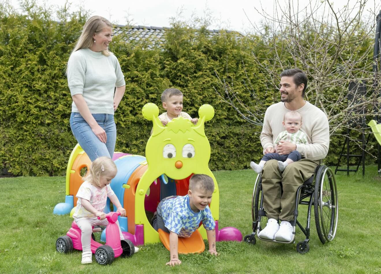 Hela familjen Schill tillsammans på gräsmattan, där de tre äldsta barnen leker med lekredskap och minstingen sitter i Jonathans knä i rullstolen.