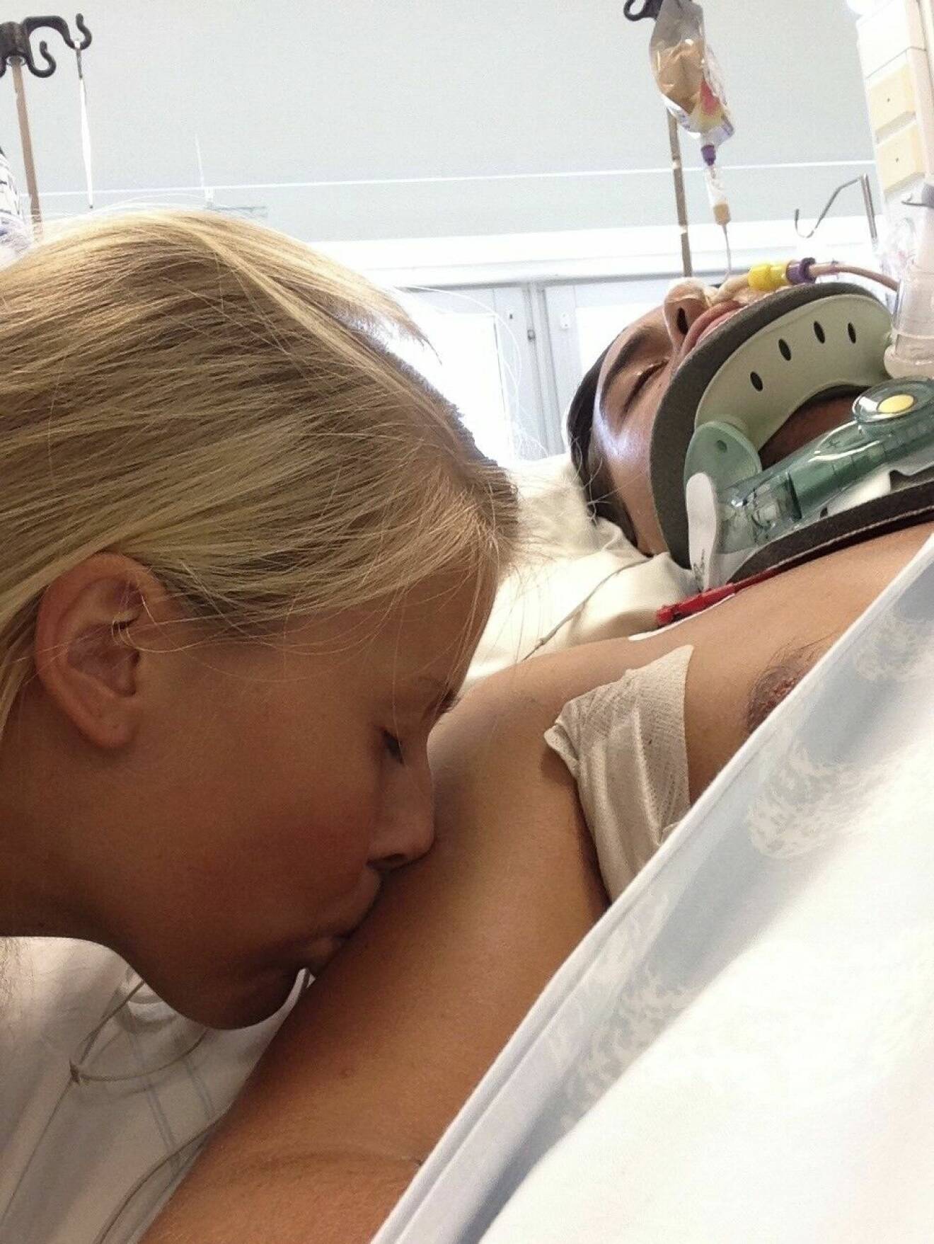 Jonathan Goding Schill ligger nedsövd på sjukhuset efter olyckan. Josefine Schill pussar på honom.