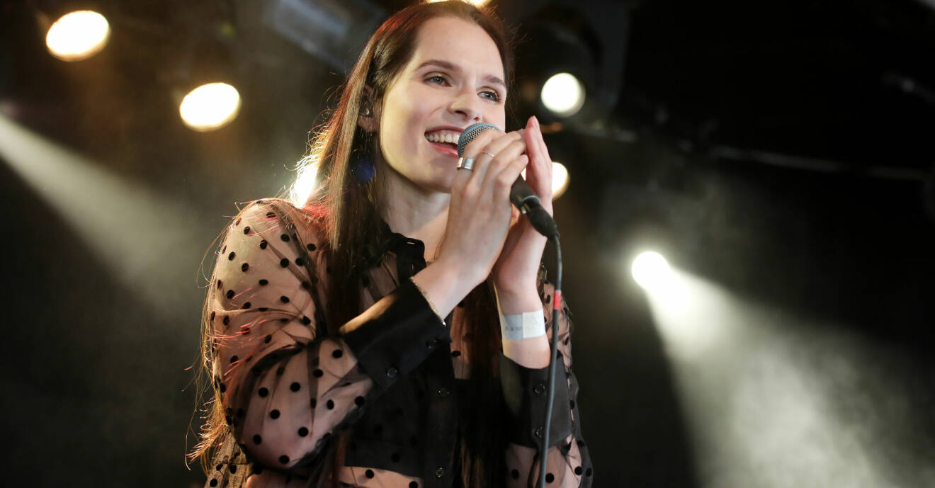 Maja Strömstedt sjunger i en mikrofon, i bakgrunden syns strålkastare. Hon har långt brunt hår och är klädd i en svart transparent blus med långa ärmar.