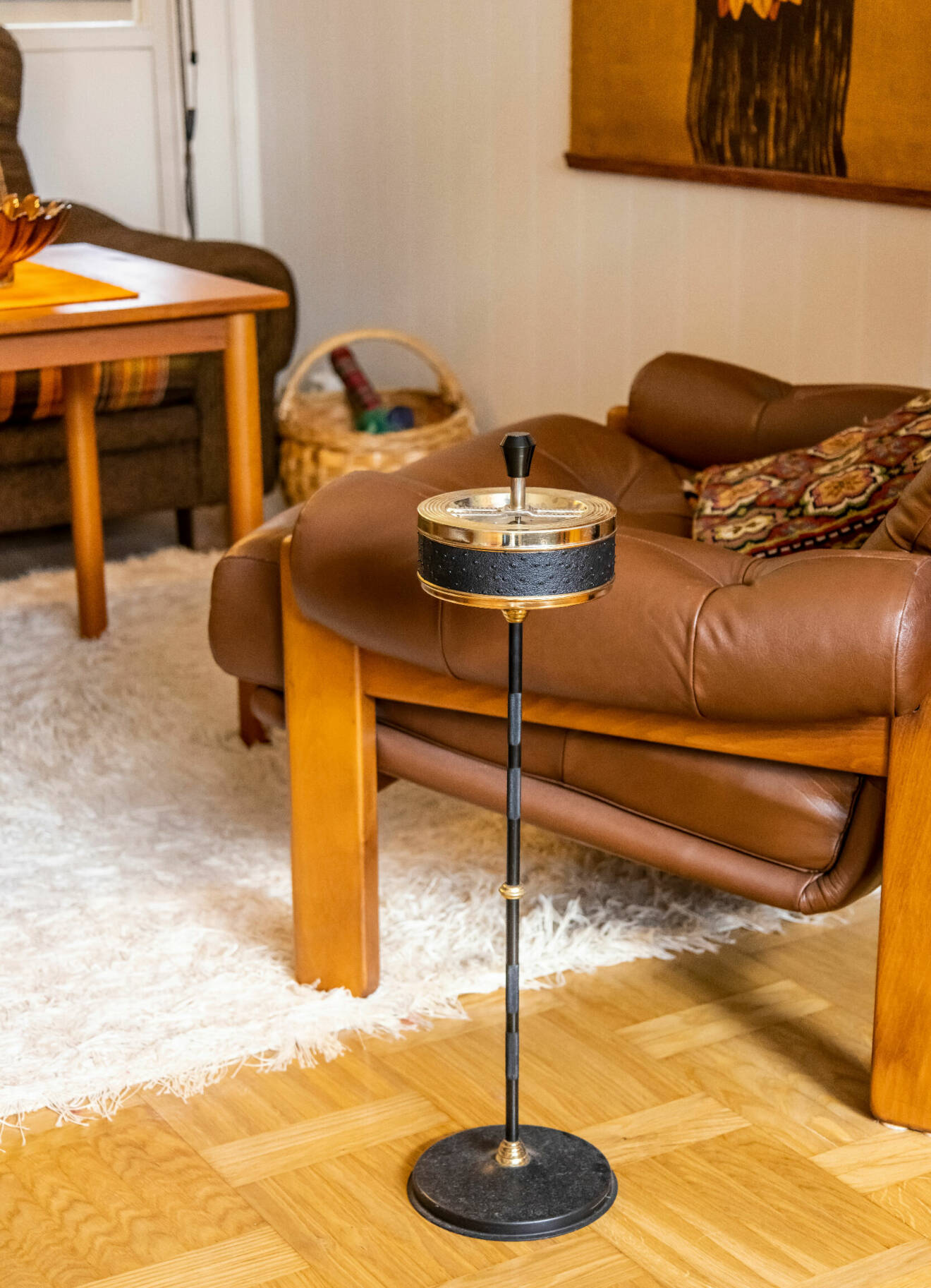 En askkopp på fot står placerad på golvet framför en brun läderfåtölj i ett vardagsrum från 70-talet.