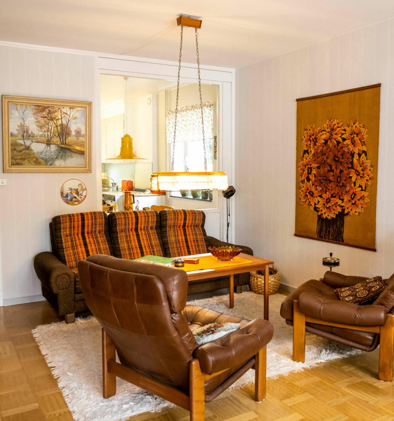 Vardagsrum i 70-talsstil. Bruna läderfåtöljer, en brunorange soffa och på väggen en bonad som också går i brunt och orange.