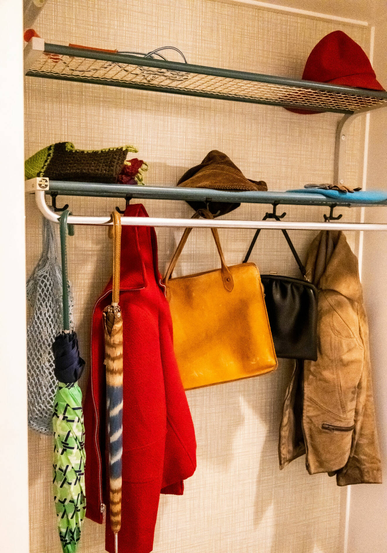En hatthylla i 70-talsstil sitter på väggen i en hall. På krokar hänger jackor och handväskor.