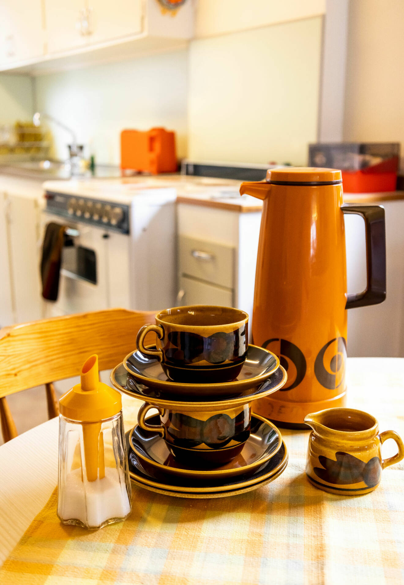 Detaljbild på ett köksbord med detaljer typiska från 70-talet. En orange kaffetermos, en brun kaffeservis och en sockerströare med orange plast.