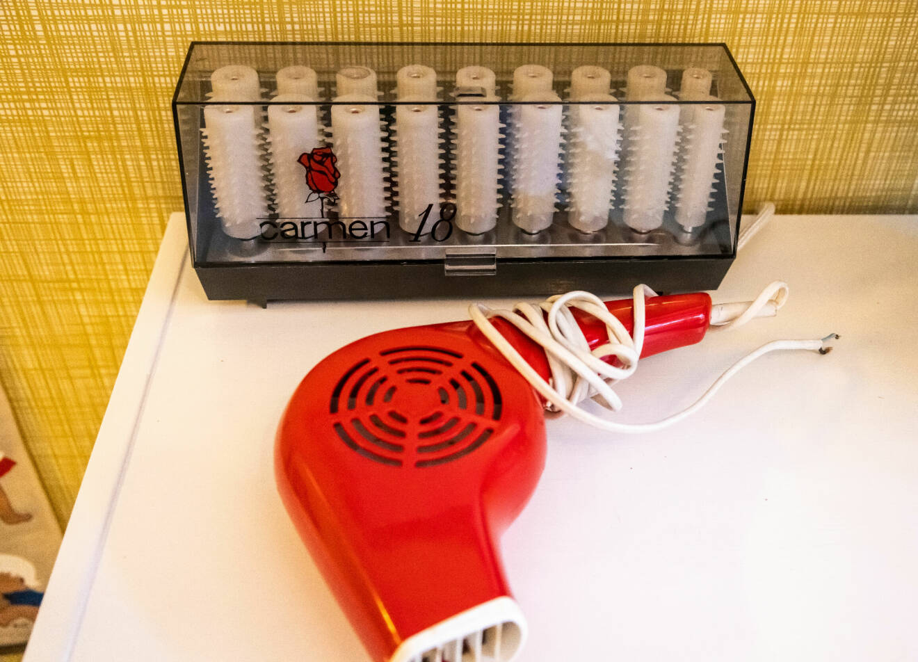 En hårfön i röd plast och en ask med värmespolar står ovanpå en tvättmaskin i en lägenhet som inretts enligt 70-talet.
