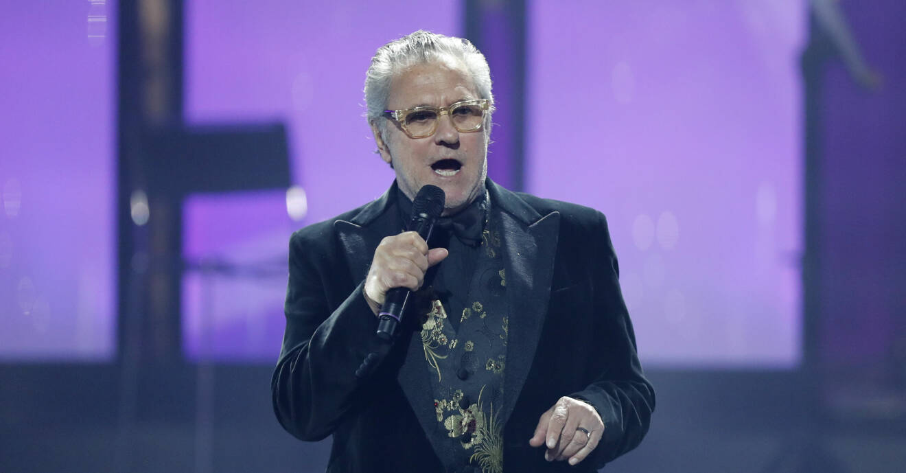 Tommy Körberg står på en scen och sjunger i en mikrofon. Han är klädd i en svart kavaj och en väst i svart och guld. Han har glasögon.