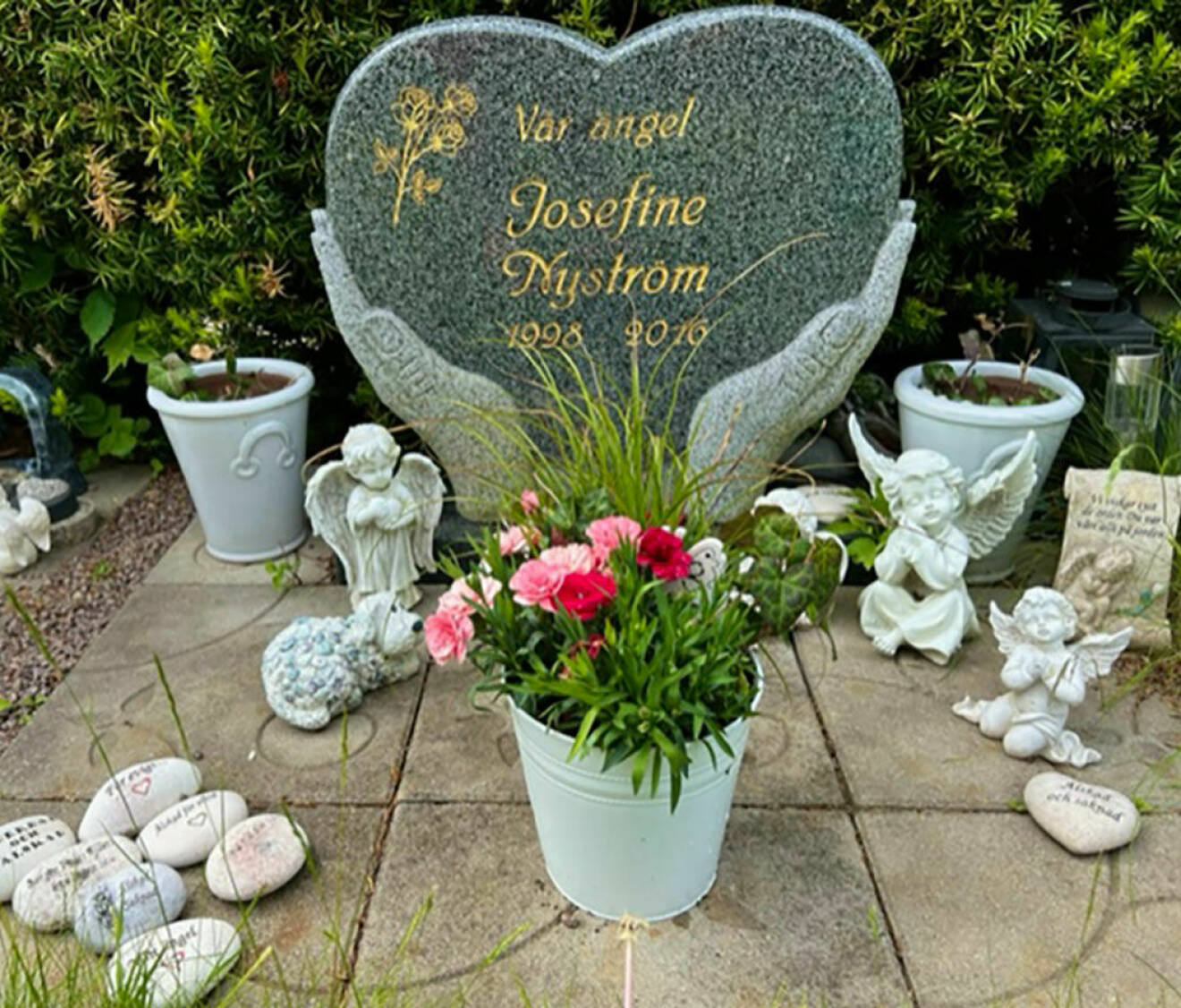 Josefine Nyströms gravsten i Vadstena. Stenen är formad som två händer som håller i ett hjärta med Josefines namn och födelsedatum. Runt gravstenen finns änglar och blommor.