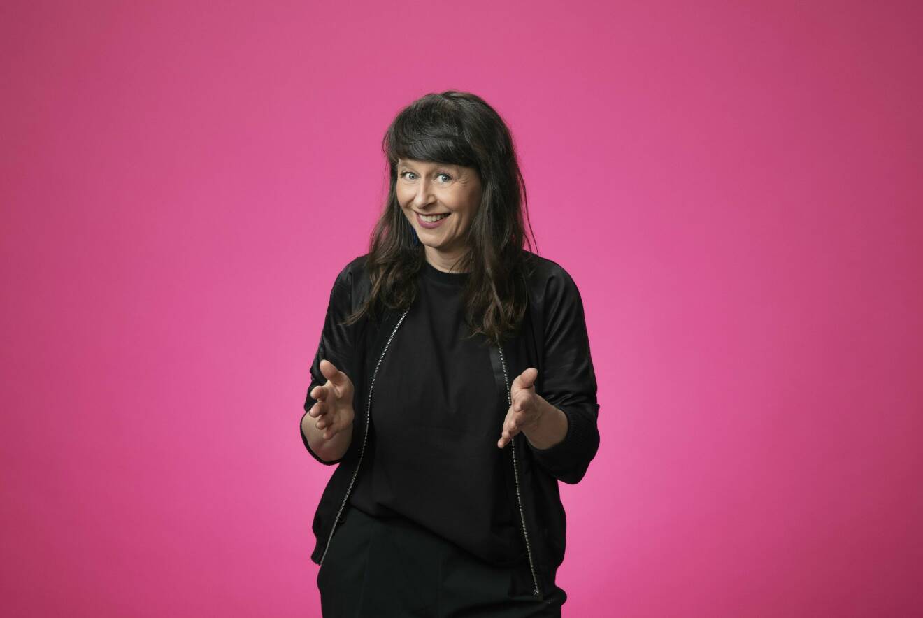 Programledaren i Melodikrysset, Anna Charlotta Gunnarson, står och gestikulerar glatt, iförd svarta kläder mot rosa bakgrund.