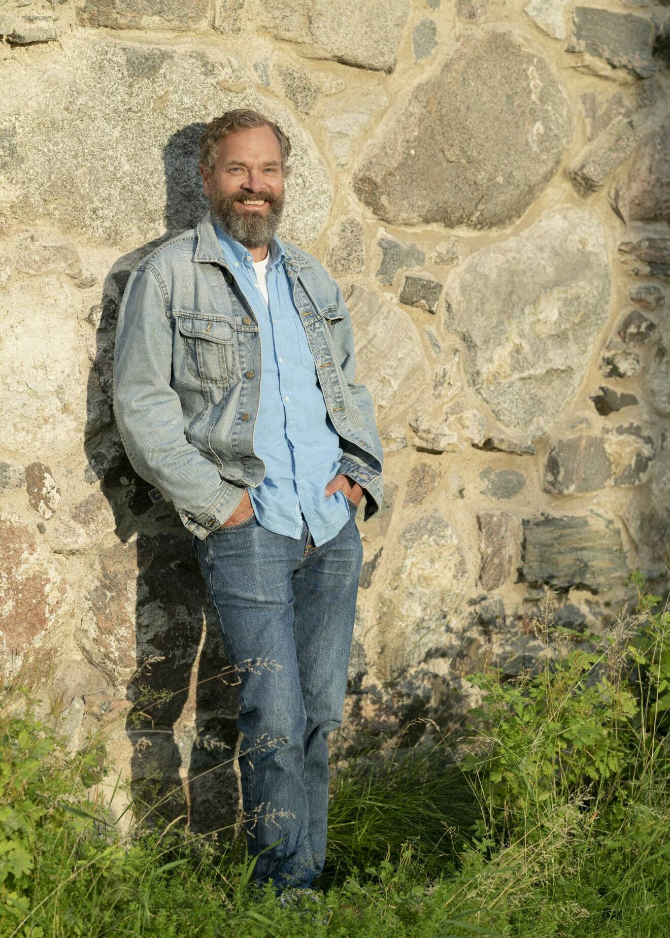 Skådespelaren Per Lasson, som ses i Tunna blå linjen, står framför en rå stenvägg, leende.