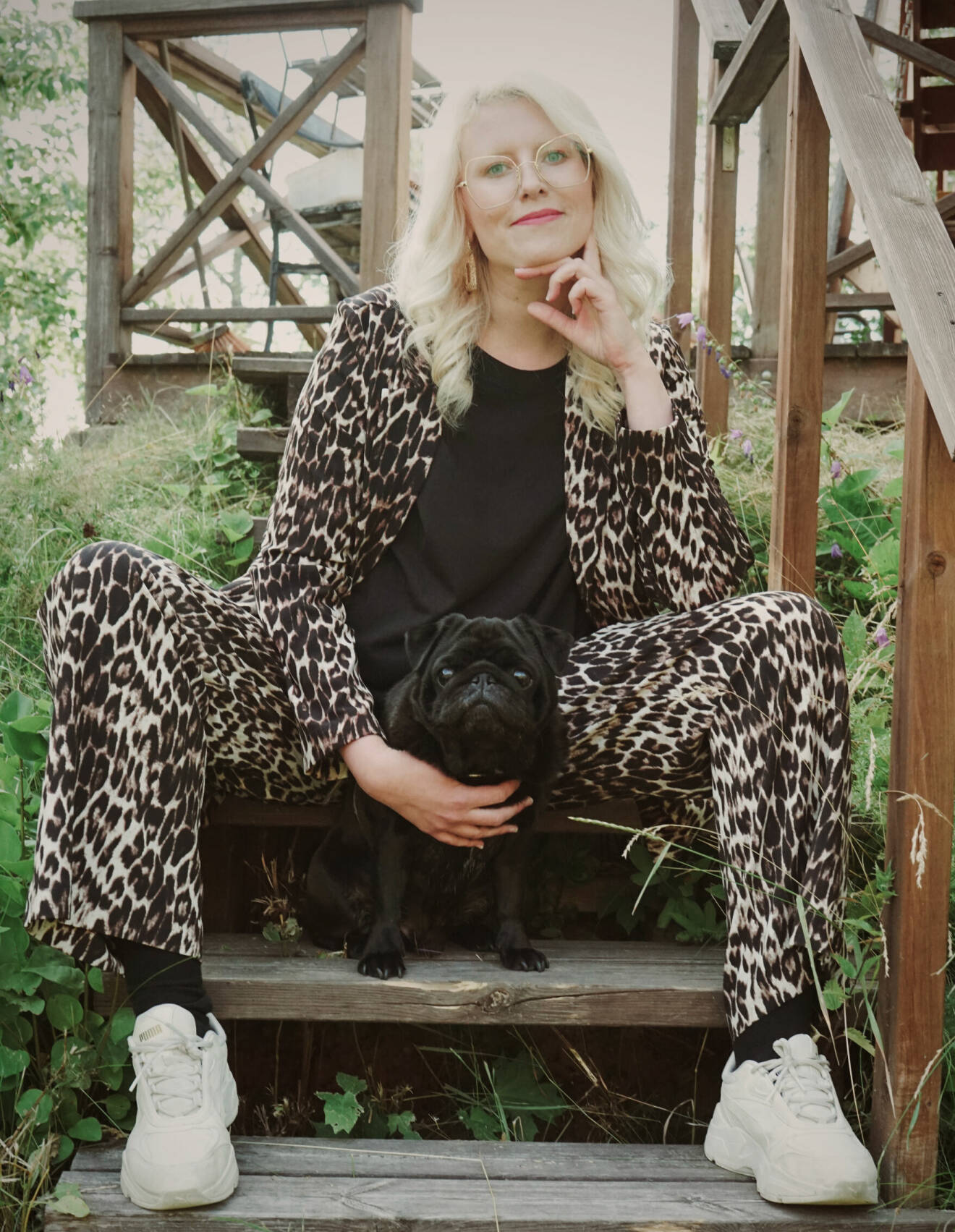 Ida Högström fotograferad utomhus, hon sitter i en trätrappa tillsammans med en svart liten mops. Hon är klädd i leopardmönstrade kläder och bär stora glasögon.