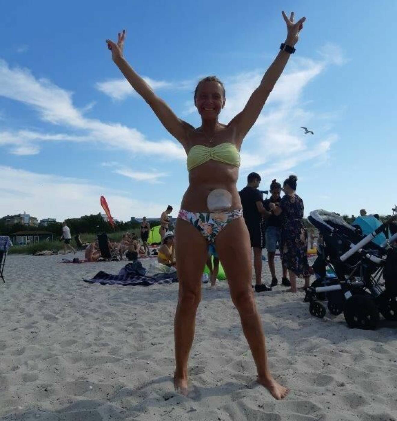 Pia Jensen står på stranden och sträcker upp händerna och ler. Hon har på sig en bikini och på magen syns hennes stomipåse.