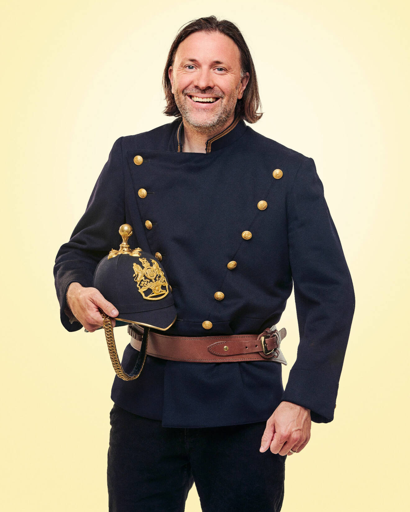 Niklas Ekstedt iklädd en gammaldags blå uniform med guldfärgade knappar och läderbälte. I ena handen håller han en tillhörande hjälm med emblem.