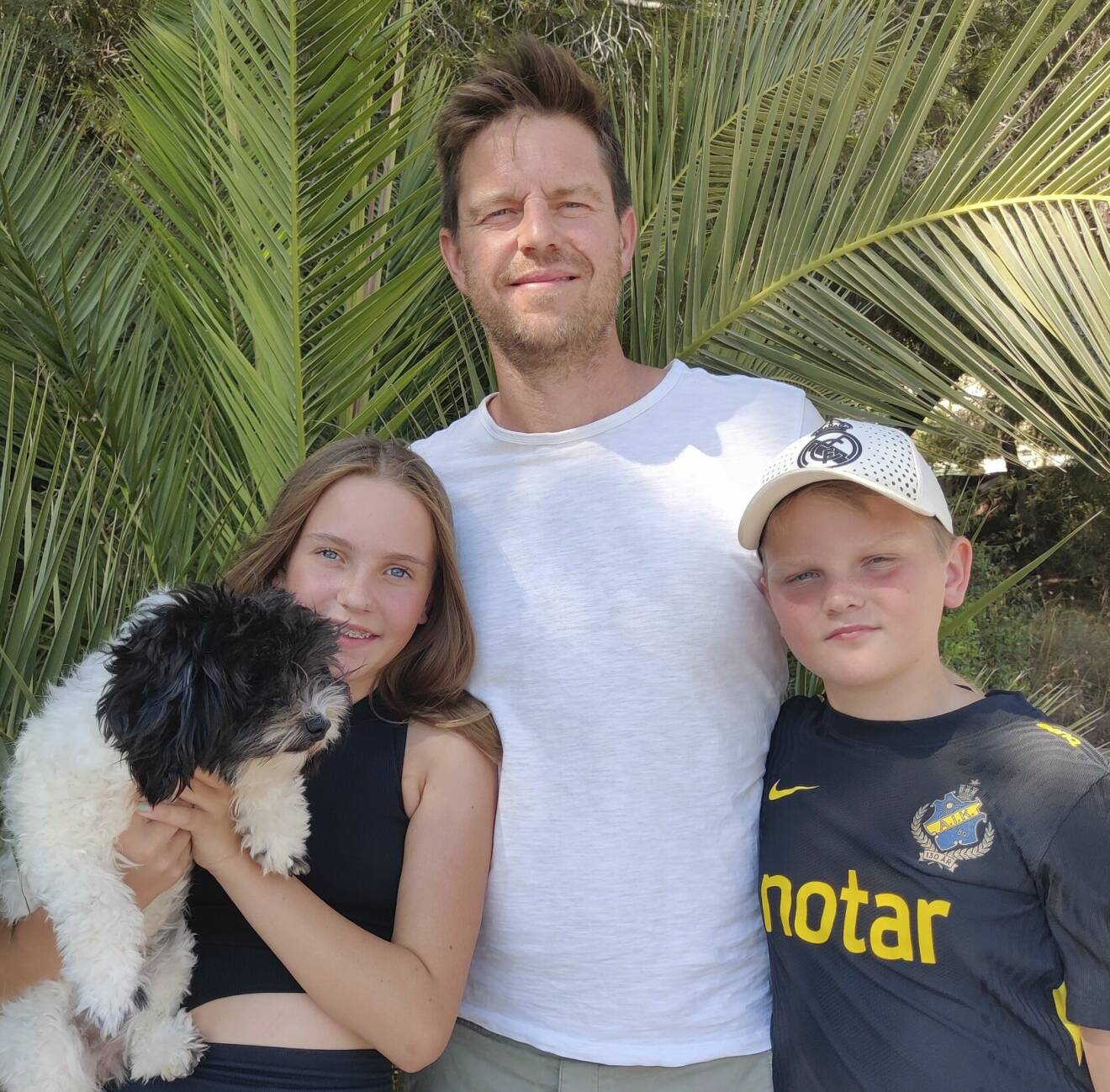 Cystisk fibros-sjuka Alice håller upp sin hund, tillsammans med sin bror och pappa under en palm.