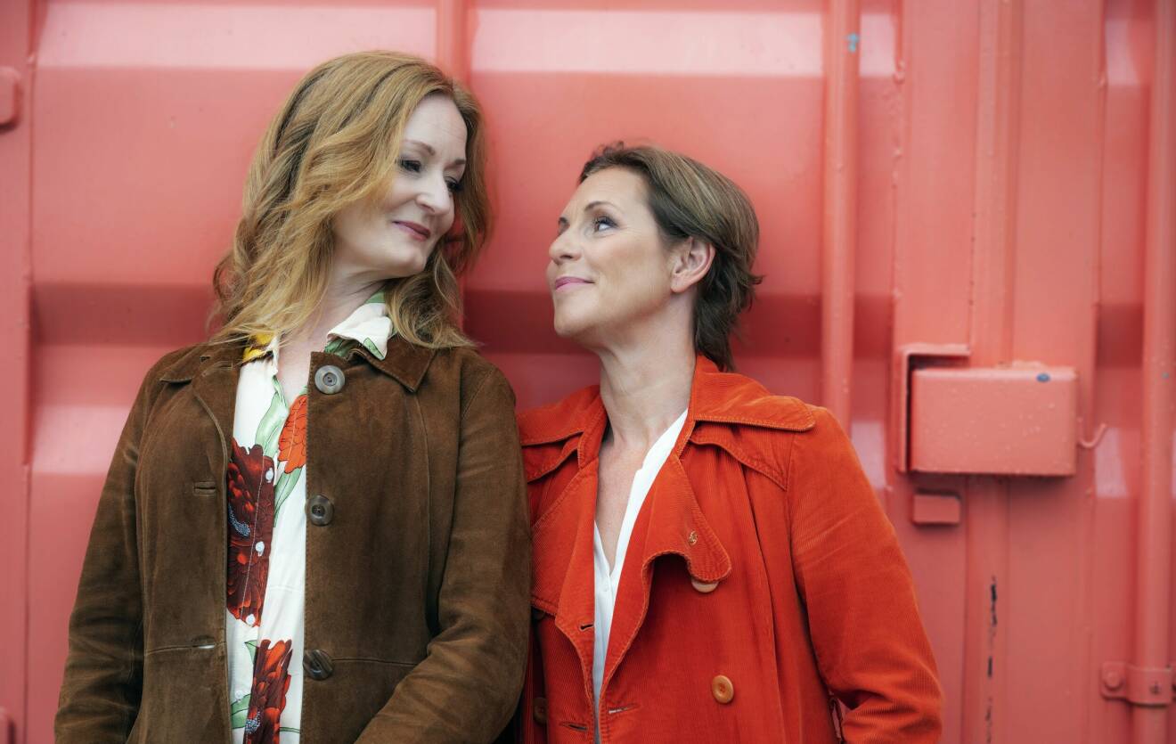 Sångerskorna Helen Sjöholm och Anna Stadling ser ömt på varandra framför en röd containervägg.