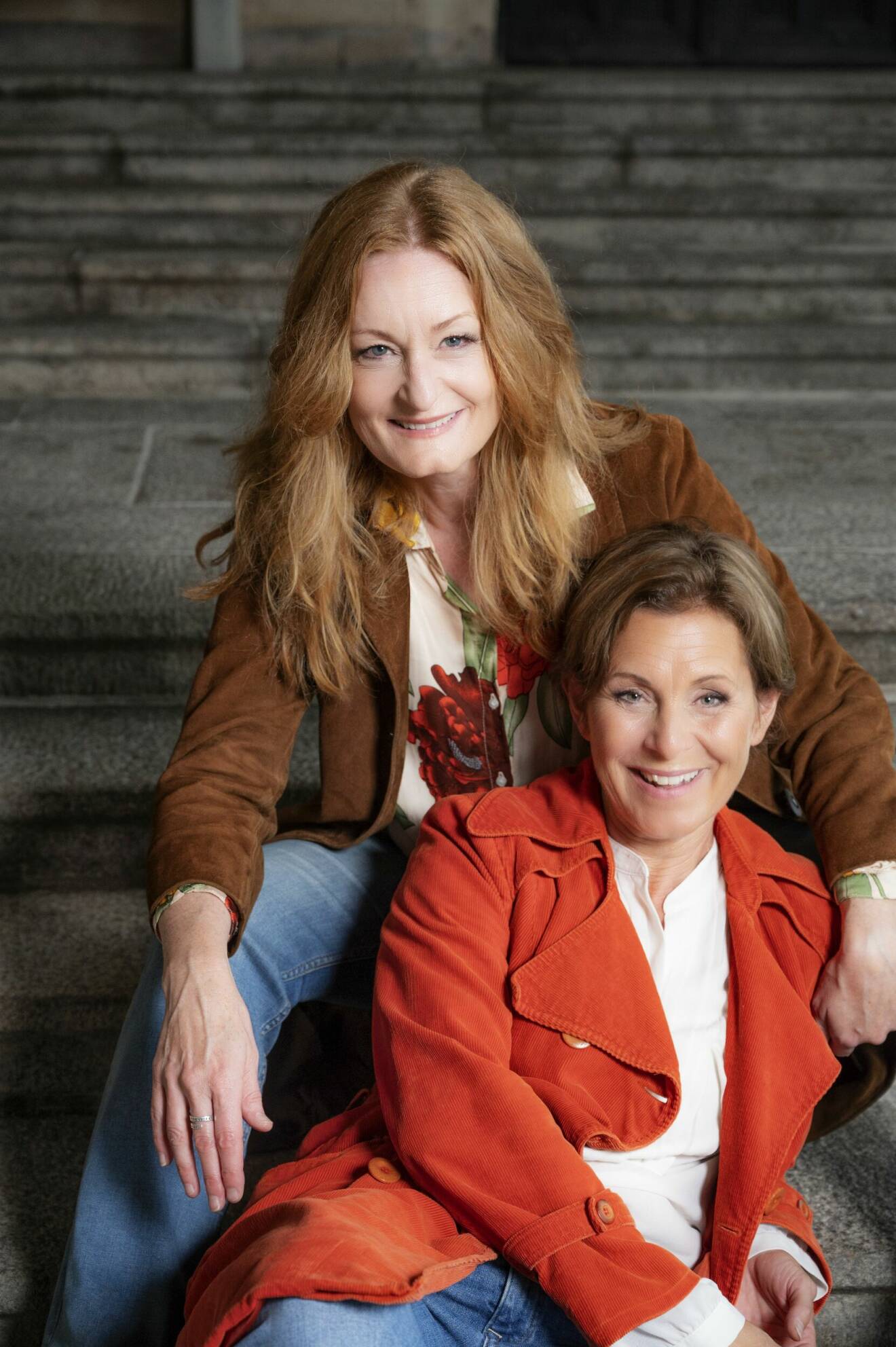 Sångerskorna Helen Sjöholm och Anna Stadling sitter i en trappa nära varandra och skrattar.