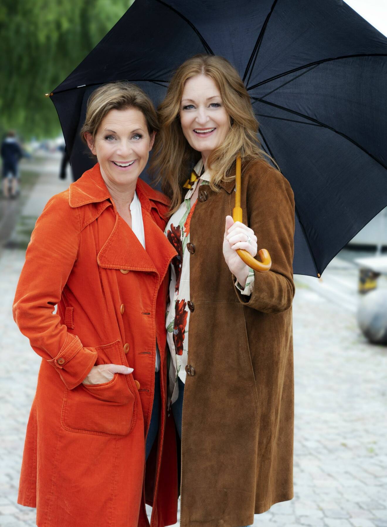 Sångerskorna Helen Sjöholm och Anna Stadling står skrattande under ett paraply.