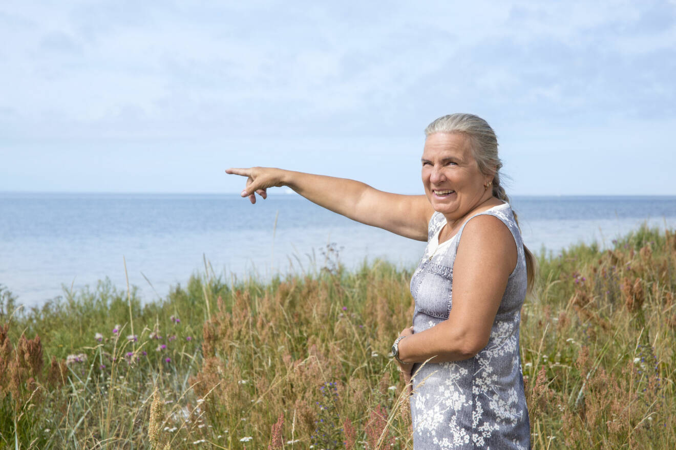 En kvinna med grått hår och blå sommarklänning skrattar och pekar ut över havet i Trelleborg.