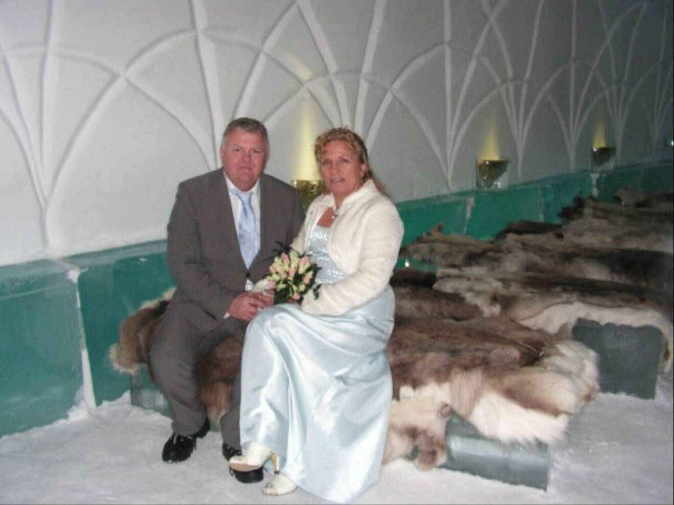 Ett medelålders par i bröllopskläder sitter på en renfäll inne i ishotellet i Jukkasjärvi.