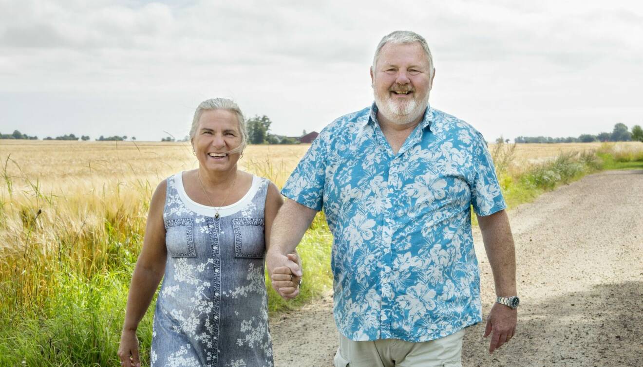 Ett par i 60-årsåldern i blå sommarkläder ler, håller varandra i handen och promenerar längs ett skånskt sädesfält.