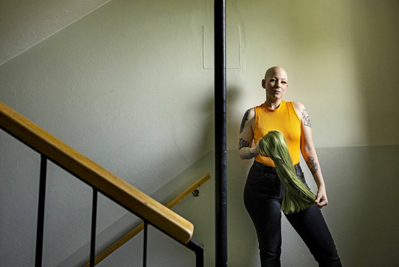 Lena Jakobsson har alopeci och kal hjässa. Hon står i trapphuset med en grön, långhårig peruk i händerna.