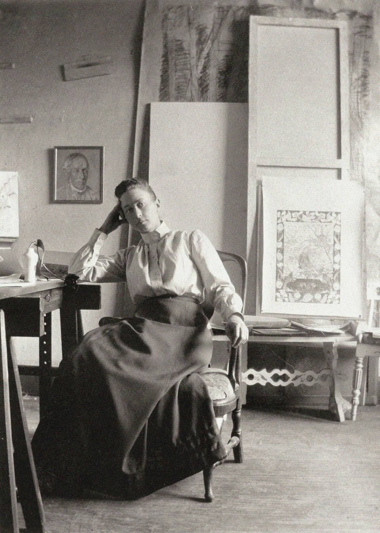Svart-vit bild på Hilma af Klint, som sitter på en stol med ett par tavlor i bakgrunden.