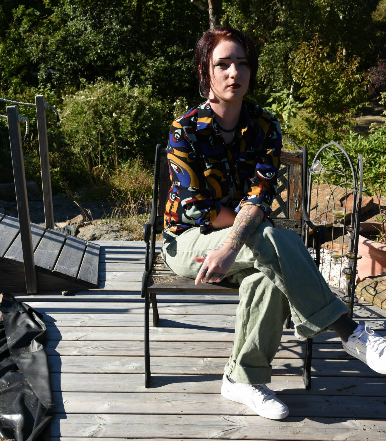Sandra Lundberg sitter på en stol utomhus på ett trädäck. Hon är klädd i en mönstrad skjorta, gröna byxor och vita sneakers.
