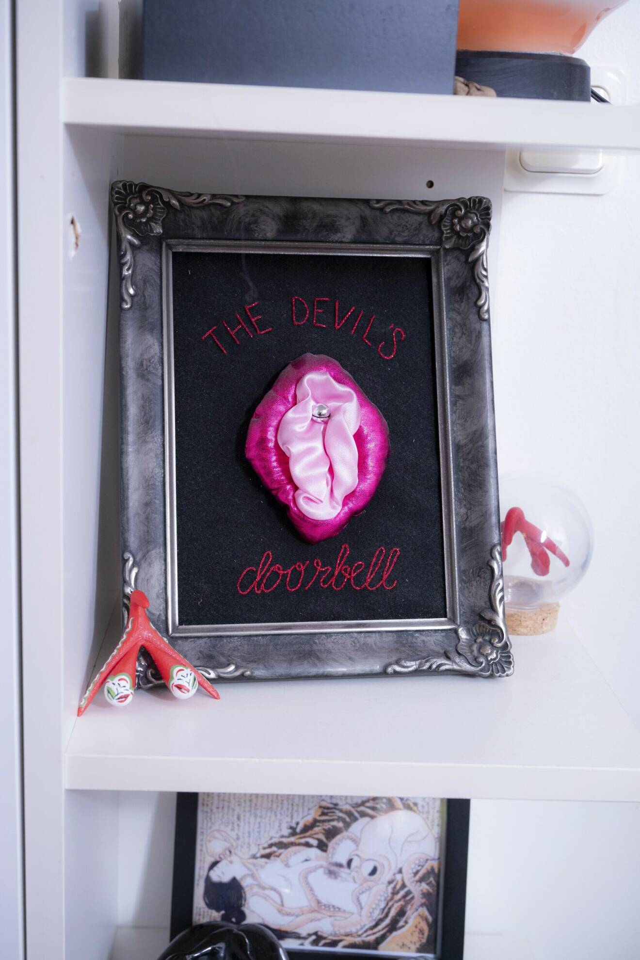En tavla med vagina och blygdläppar i tyg och klitoris som en silvrig knopp. Runt motivet finns texten "The devil's doorbell".