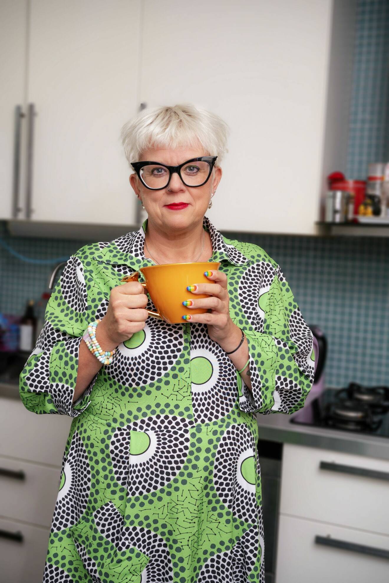 Sexologen Suzann Larsdotter står hemma i köket med en orange tekopp i händerna. Hennes naglar är målade som Pride-flaggan.