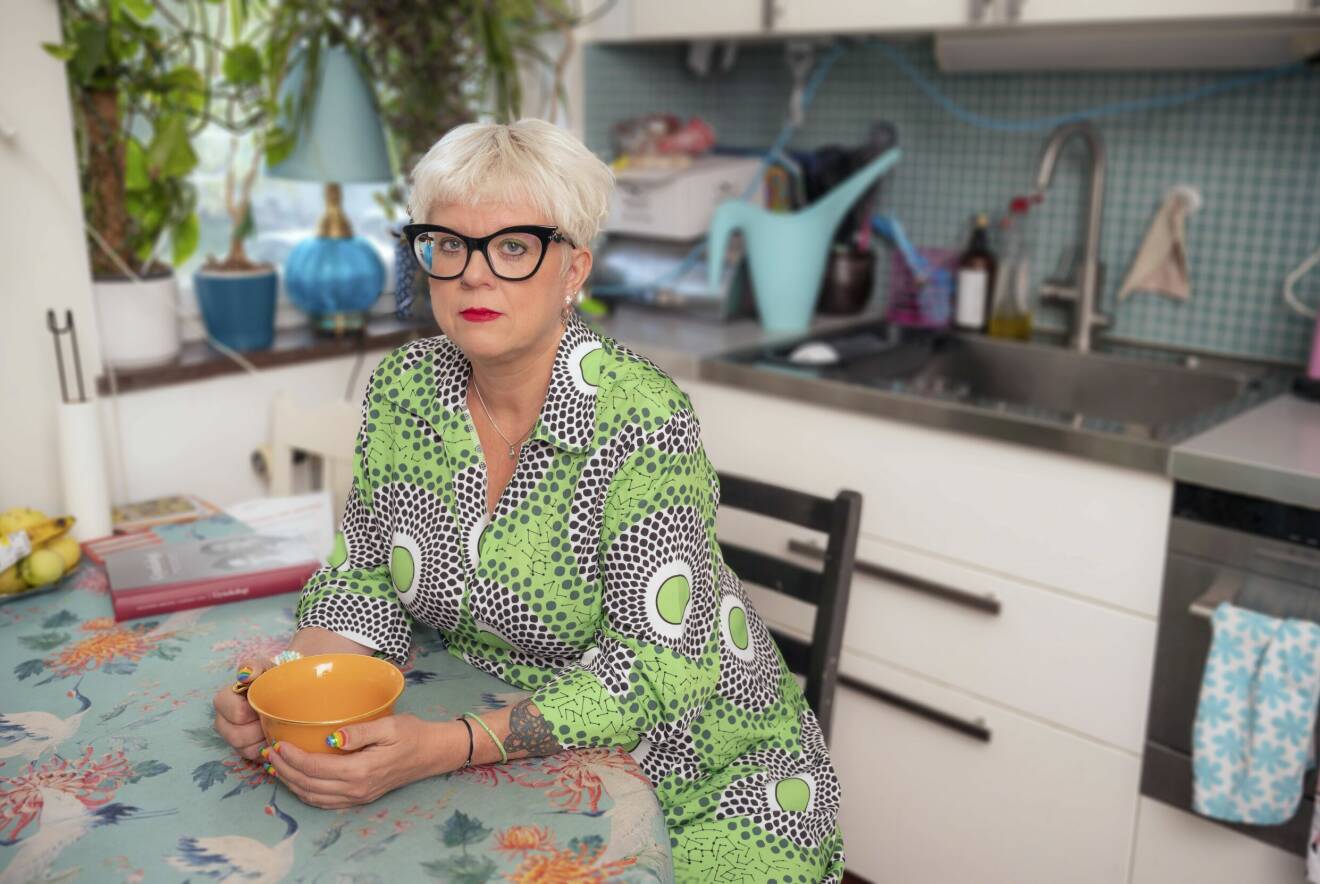 Sexologen Suzann Larsdotter sitter hemma vid köksbordet med en orange tekopp i händerna. Naglarna är målade som Pride-flaggan.