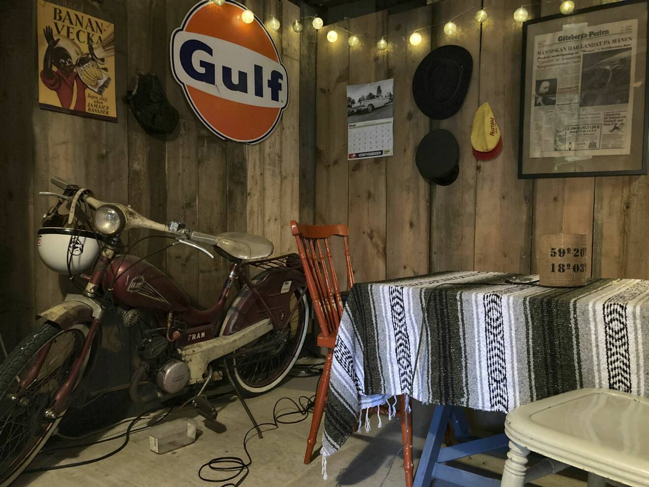 En samling retroprylar i hemmiljö, däribland en Gulf-skylt, en gammal moped, pinnstol, vävd duk och tidningsklipp på väggen.