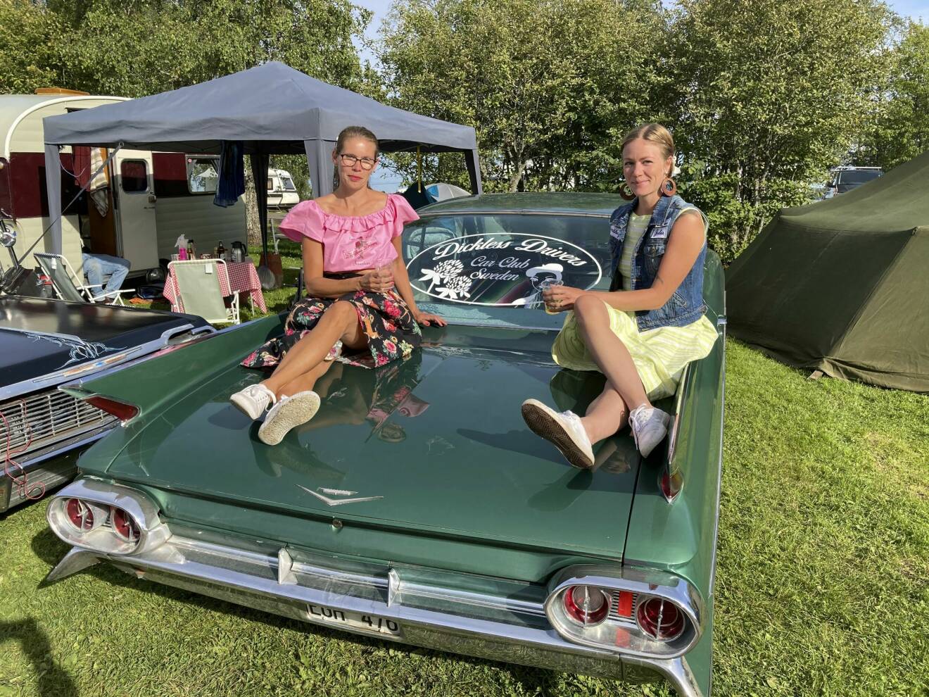 Annelie Sundberg och Julia Lind, i färgglada kjolar, sitter uppkrupna på veteranbilen med Dickless Drivers emblem på bakrutan mellan sig.