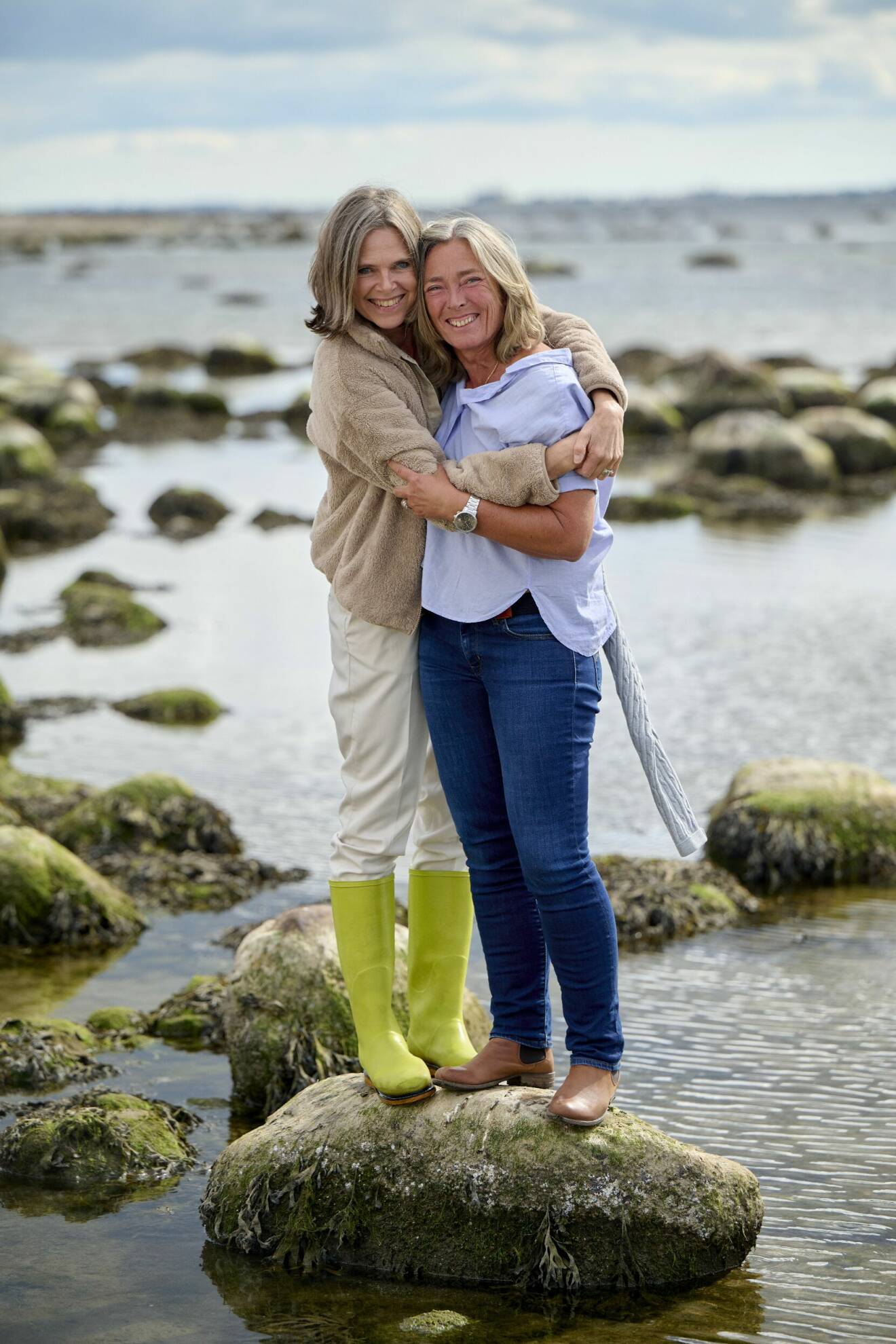 Birgitta Hågemark Olshov håller om Annika Erlandsson Olshov där de leende står tillsammans på en stor sten i vattnet nära strandkanten i Viken vid Öresund.