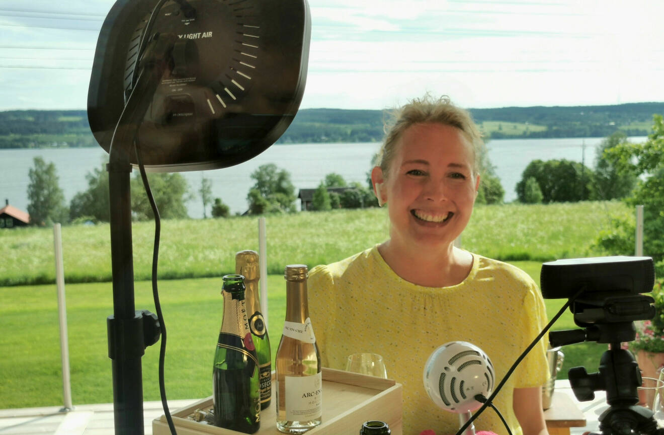 Camilla sitter utomhus med sin inspelningsutrustning för en digital vinundervisning framför sig. Grönt landskap syns i bakgrunden.
