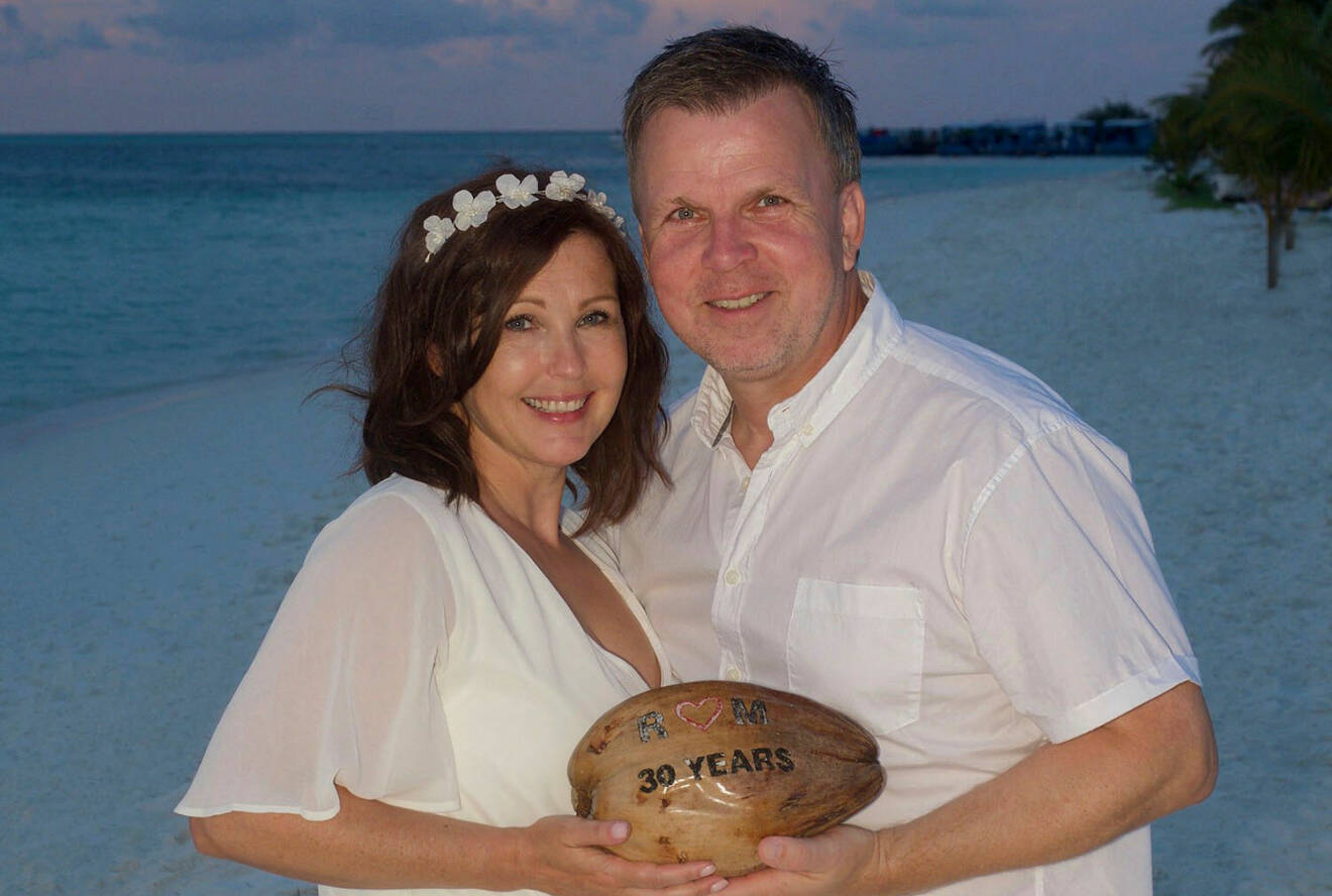 Marie och Richard står på en sandstrand i vita kläder. De ler och håller i en stor sten där det står "30 år" på.